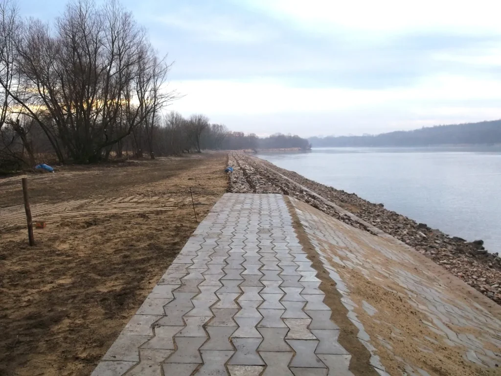 Ukończony zjazd techniczny z betonowej kostki sześciokątnej (trylinki) prowadzący do opaski brzegowej nad Wisłą w Warszawie. Po prawej stronie widoczna skarpa zabezpieczona narzutem kamiennym, w tle rzeka i drzewa.