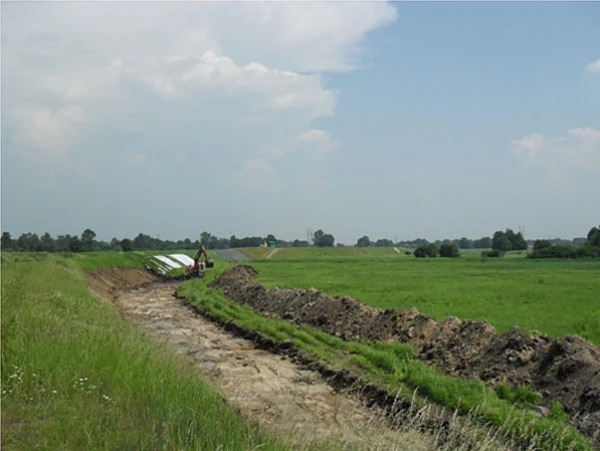 Prace ziemne zmechanizowane przy budowie wałów przeciwpowodziowych w widłach Wisły i Soły. Koparka przygotowuje podłoże pod nasyp, widoczna hałda ziemi oraz rozległe tereny zielone pod błękitnym niebem.