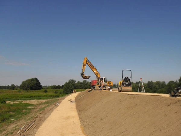 Ciężki sprzęt budowlany podczas formowania wału przeciwpowodziowego rzek Wisły i Soły. Na szczycie nasypu pracuje koparka gąsienicowa oraz walec drogowy zagęszczający nawierzchnię korony wału.