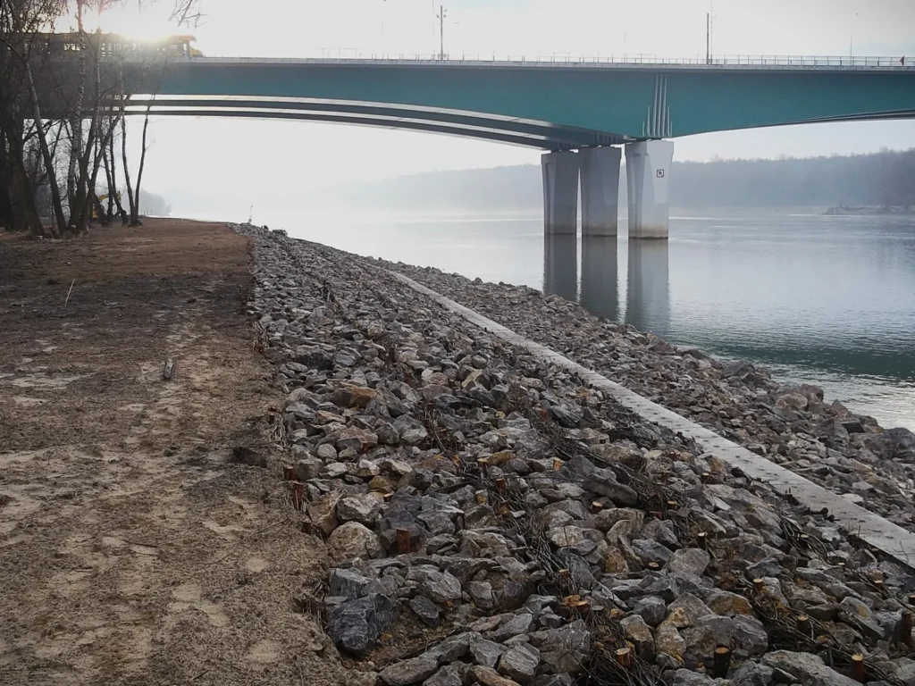 Panorama opaski brzegowej w Warszawie z Mostem Marii Skłodowskiej-Curie w tle. Na pierwszym planie umocnienia z kamienia i faszyny, w oddali filary mostu oraz przejeżdżający tramwaj na tle mglistego krajobrazu.