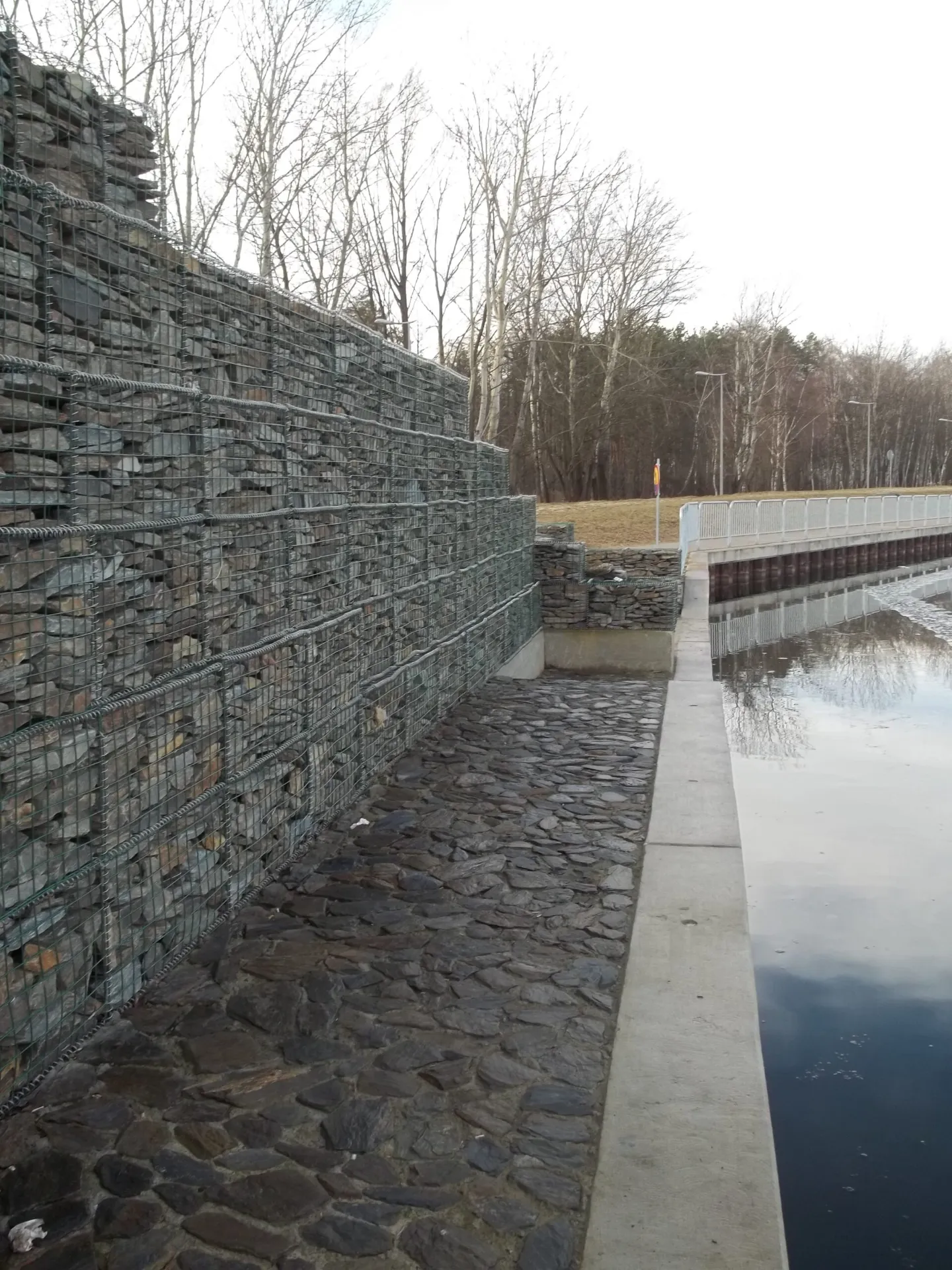 Wysoki mur oporowy wykonany z gabionów (koszy stalowych wypełnionych kamieniami) chroniący brzeg Portu Czerniakowskiego. Obok betonowy chodnik serwisowy i barierka, wzdłuż których rozciąga się tafla kanału portowego.