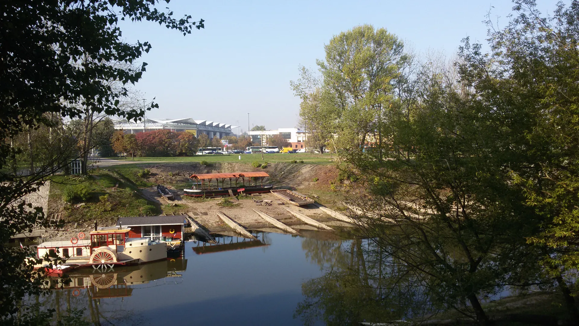 Archiwalny widok na teren dawnej stoczni w Porcie Czerniakowskim przed modernizacją. Na brzegu widoczne stare tory wyciągowe, drewniane zadaszenie i cumujące jednostki pływające w otoczeniu gęstej zieleni.