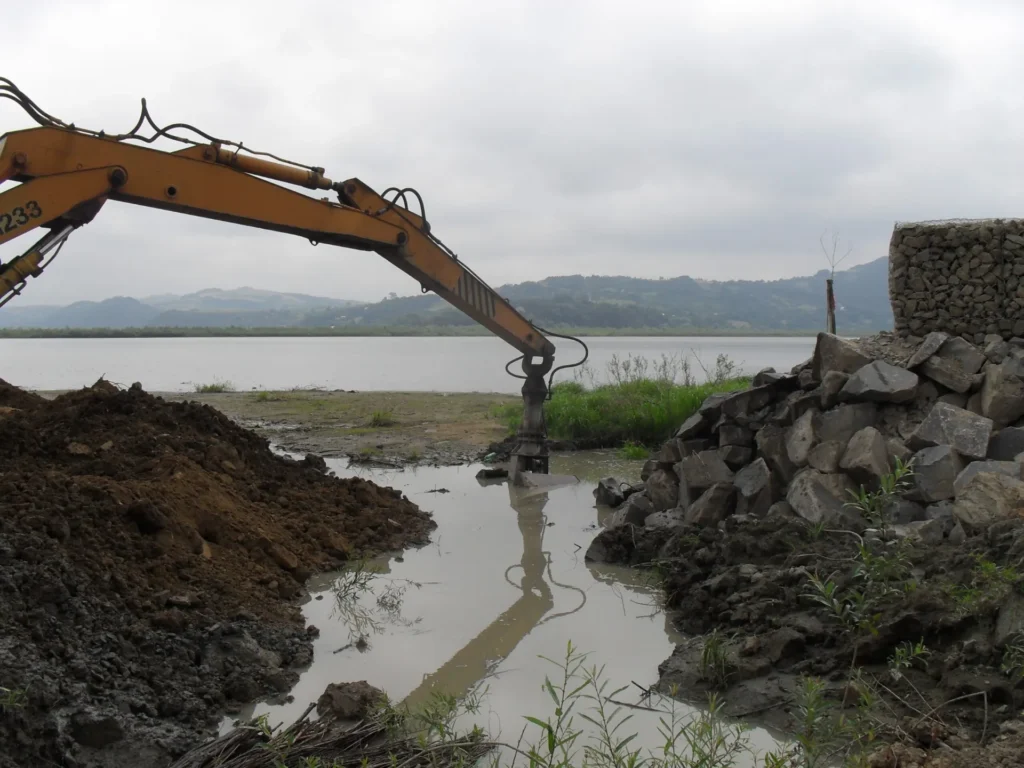 Ramie koparki pracującej przy brzegu zbiornika Czchów-Rożnów. Maszyna operuje przy pryzmach ziemi i kamieniach, obok widać kosz gabionowy wypełniony skałami. W tle zamglony krajobraz pagórków.