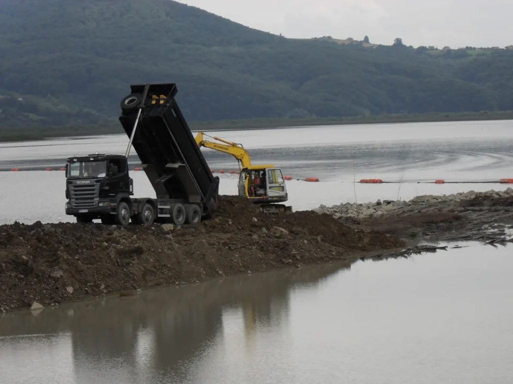 Ciężarówka wywrotka marki Scania wysypuje urobek na wąskim nasypie ziemnym wzdłuż brzegu zbiornika. Obok pracuje koparka gąsienicowa marki Hyundai. W tle widać wzgórza pokryte lasem i linię boi na wodzie.