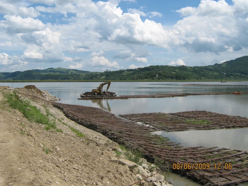 Prace przy umacnianiu brzegu zbiornika w Tęgoborzu. Na pierwszym planie materace faszynowe ułożone na wodzie, w oddali koparka na barży (pontonie) oraz linia brzegowa z widocznymi wzniesieniami.