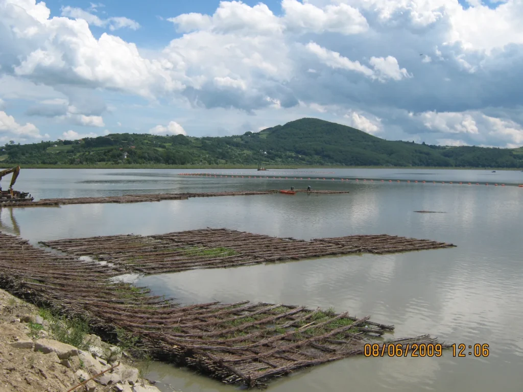 Szerokie ujęcie zbiornika wodnego z lotu ptaka (z poziomu brzegu) prezentujące konstrukcje z faszyny służące do rekultywacji i ochrony brzegów przed erozją. Na wodzie widoczna łódź z pracownikami oraz pomarańczowe boje.
