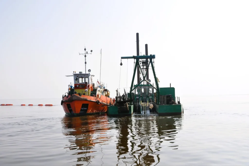 Prace przy udrożnieniu Przekopu Wisły w Gdańsku. Na tafli wody widoczne jednostki pływające. W tle rozciąga się linia boi wyznaczająca obszar roboczy robót czerpalnych.